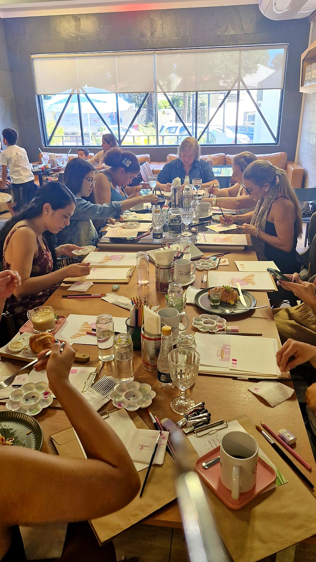 Grupo de mulheres pintando aquarela em uma mesa de café, com xícaras de café e materiais artísticos espalhados